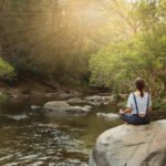 Woman Meditating On Rock By River In Forest