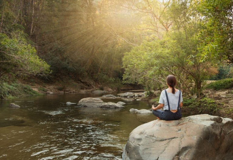 Woman Meditating On Rock By River In Forest