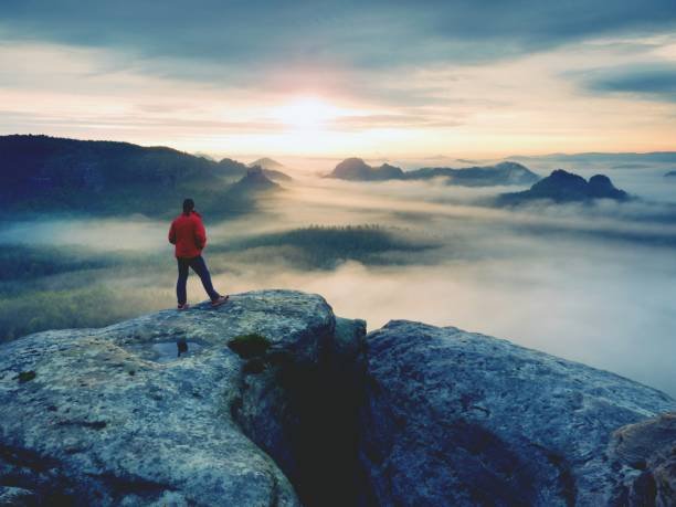 Hiker in red jacket enjoy view over clouds. Mountain rocky peak above valley.