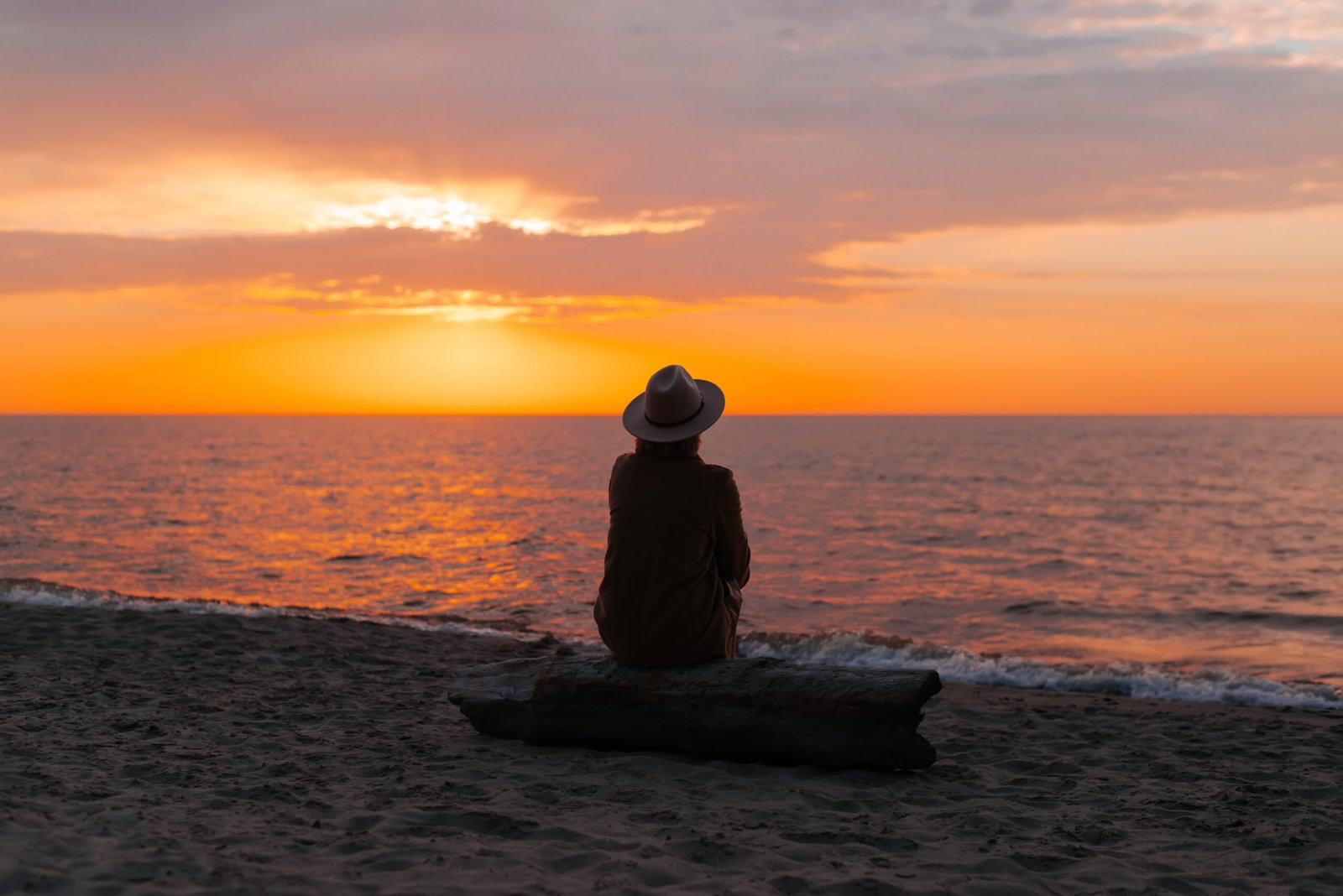 Back view of lonely adult thoughtful woman in coat and hat sitting on og on seashore and looking at sea sunset, mood. Unrecognizable pensive senior lady alone in nature in evening