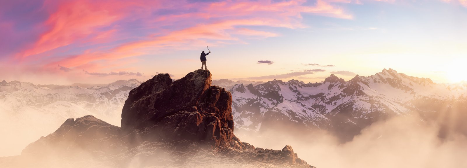 Adventurous Man Hiker standing on top of icy peak with rocky mountains in background.