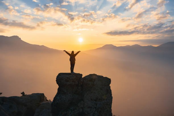 Silhouette of sporty girl standing on mountain top with raised arms at sunset