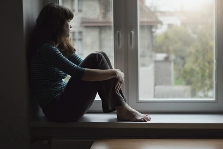 Sad young woman sitting on the window at home isolated, watching out. Coronavirus quarantine concept.