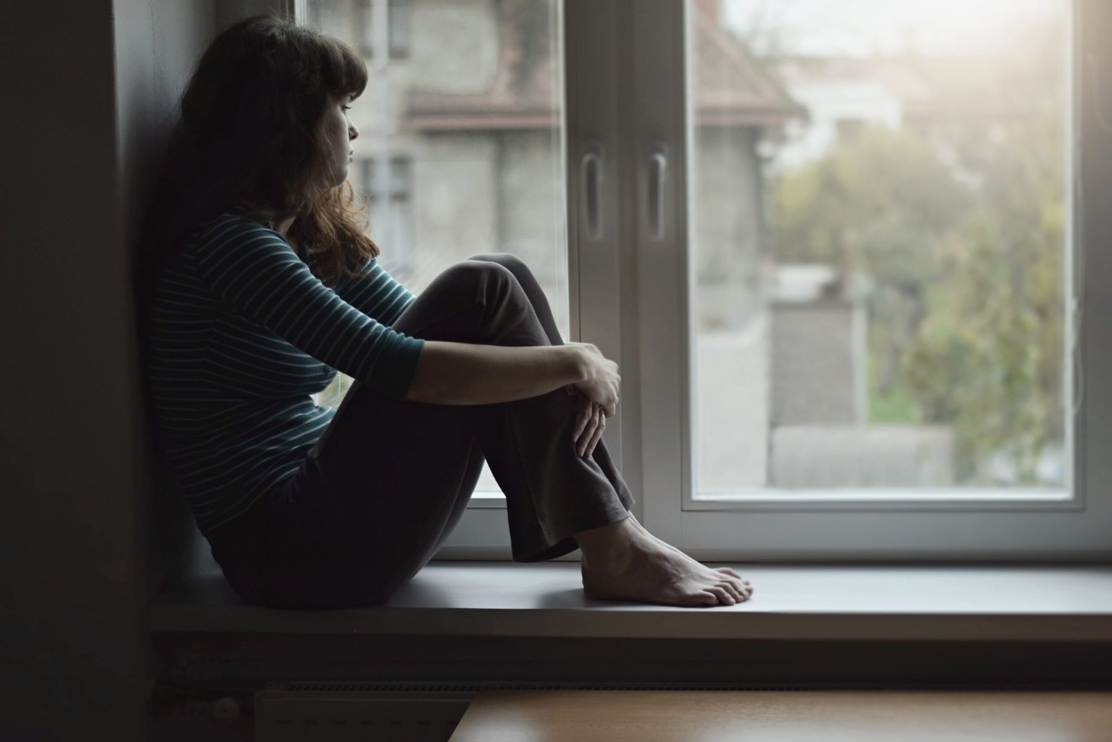 Sad young woman sitting on the window at home isolated, watching out. Coronavirus quarantine concept.