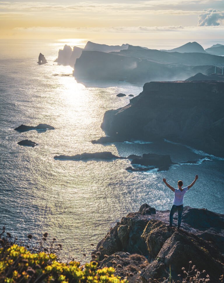 Male Tourist reaches his arms in the air at sunrise from a vantage point on a steep cliff overlooking the sea and along the rugged foothills of Madeira's coastline with wind turbines. Ponta do Bode, Madeira Island, Portugal, Europe.