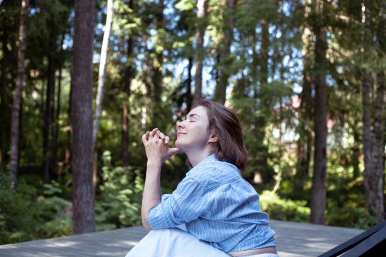 A woman sits peacefully, hands clasped, with a content expression on her face. Sunlight filters through the trees, creating a tranquil atmosphere in the forest