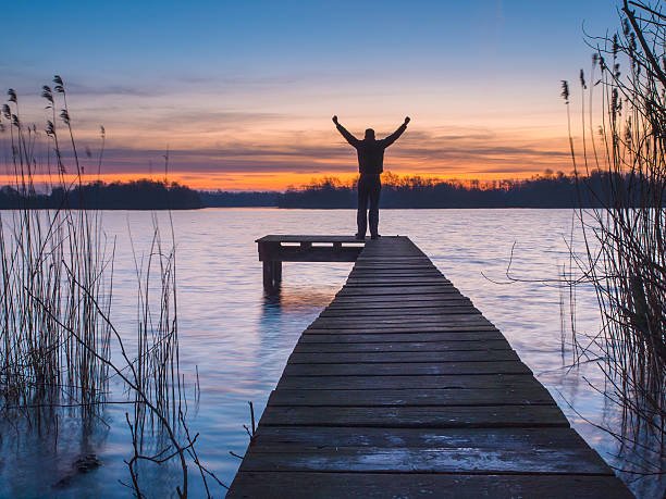 Man on a jetty raising arms during sunset as a sign of achievement