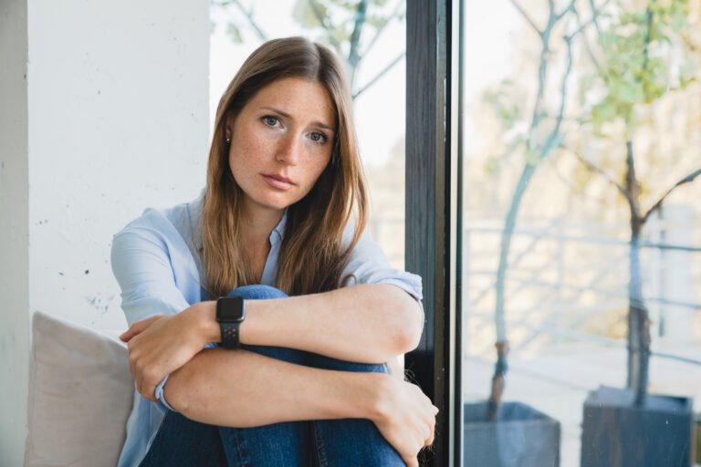 Depressed young woman thinking about breakup sitting by the window alone at home.