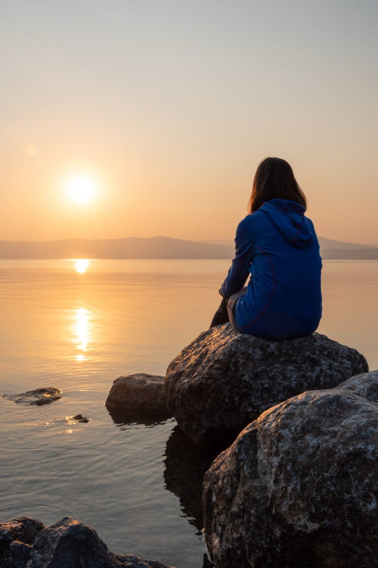 Girl looking sunset on sea, lake. Woman sitting on rocks. Lonely person. Girl alone outside. Vacation on sea. Relax sunset silhouette