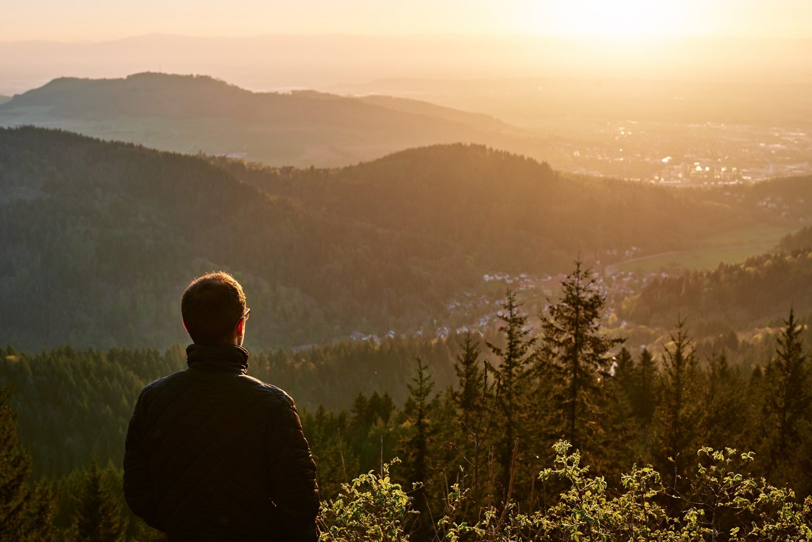 One man looking a sunrise on a peak of a mountain. Back of the man looking a forest. Handsome male in the nature. Sunrise from the top of a mountain with a town and a man.