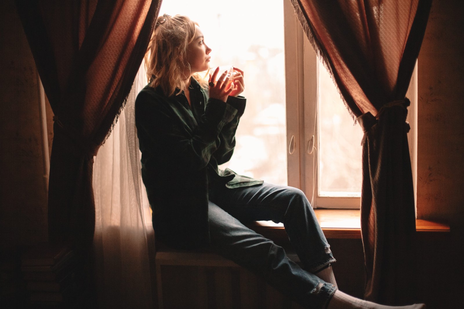 Young woman drinking tea sitting on windowsill at home
