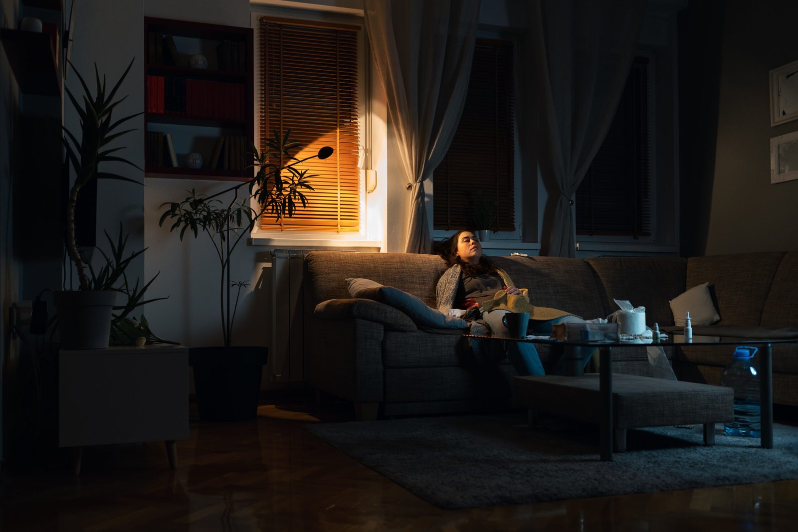 Depressed young Caucasian woman covered blanket sits on sofa in dark living room with electric light. Copy space. Concept of psychology illness and treatment
