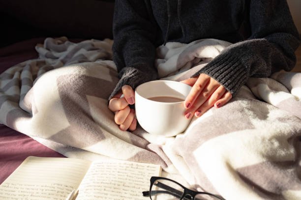 Female hands holding steaming cup of hot cocoa reading in bed