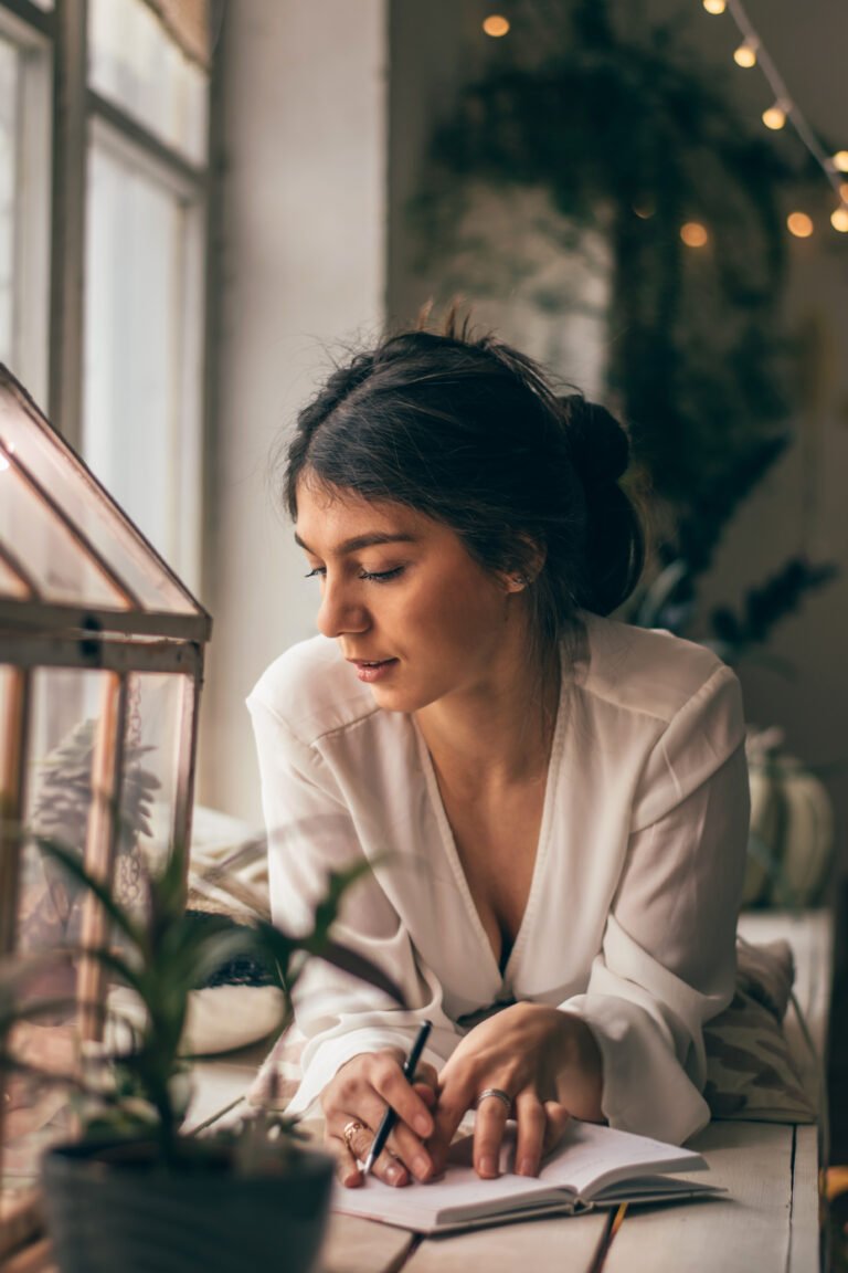 A young attractive dark-haired woman is lying on the windowsill in a greenhouse room.A smiling woman writes in a notebook