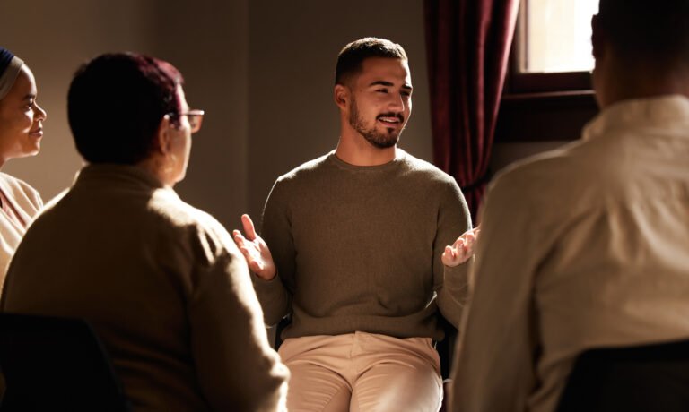 Support, smile and man sharing in group therapy with understanding, feelings and talking in session. Mental health, addiction or depression, men and women with therapist sitting together for healing.