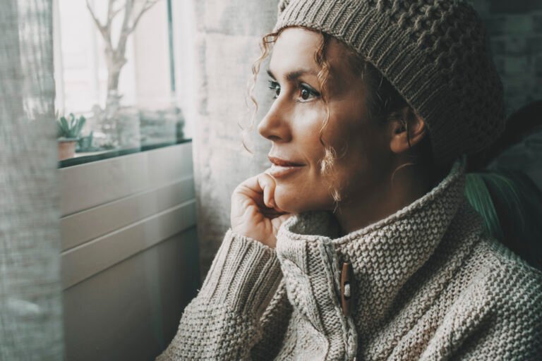 Side portrait of serene female looking outside the window at home in winter season Cold house temperature. One woman wearing knit hat inside apartment. Relax and feeling expression lady leisure