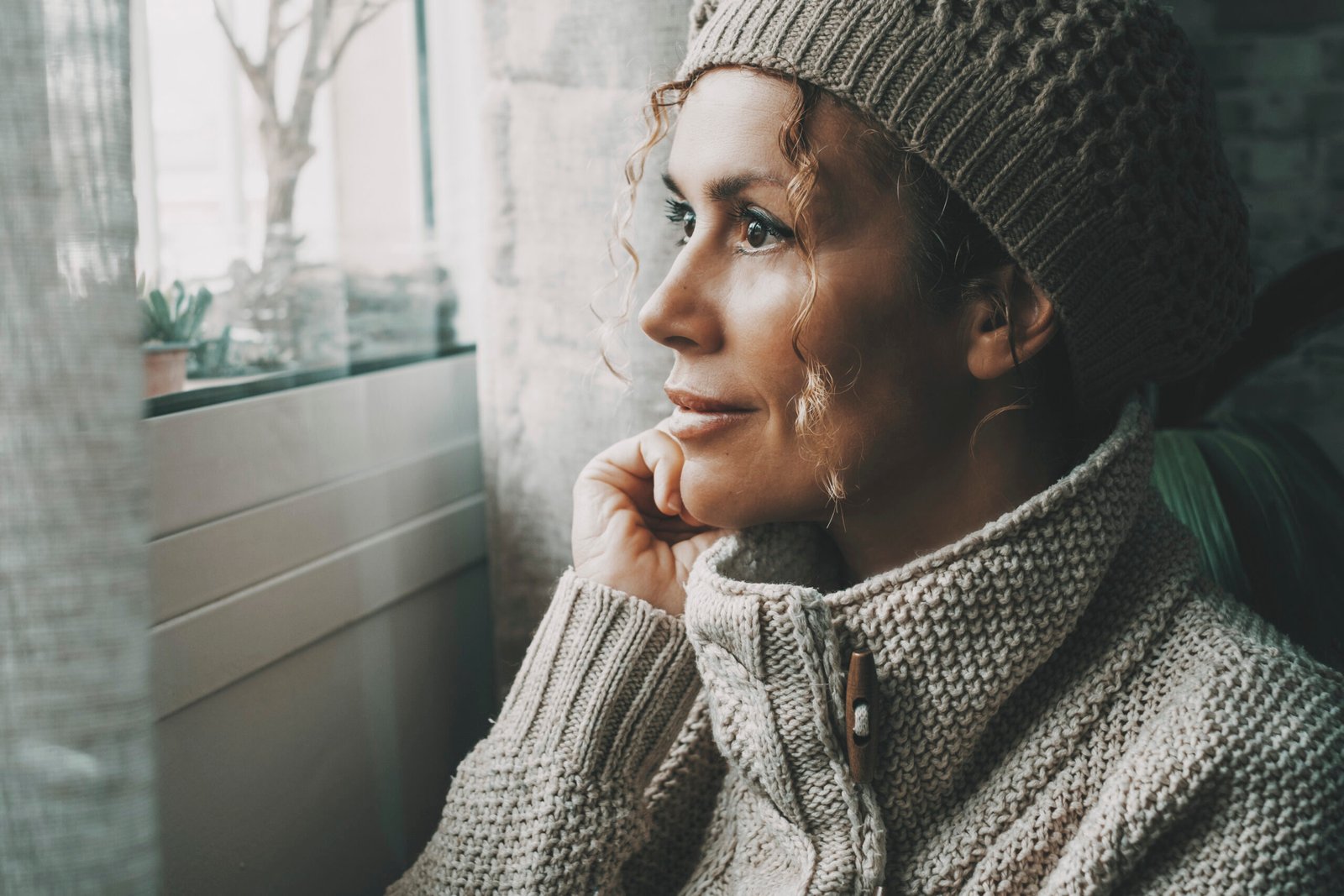 Side portrait of serene female looking outside the window at home in winter season Cold house temperature. One woman wearing knit hat inside apartment. Relax and feeling expression lady leisure