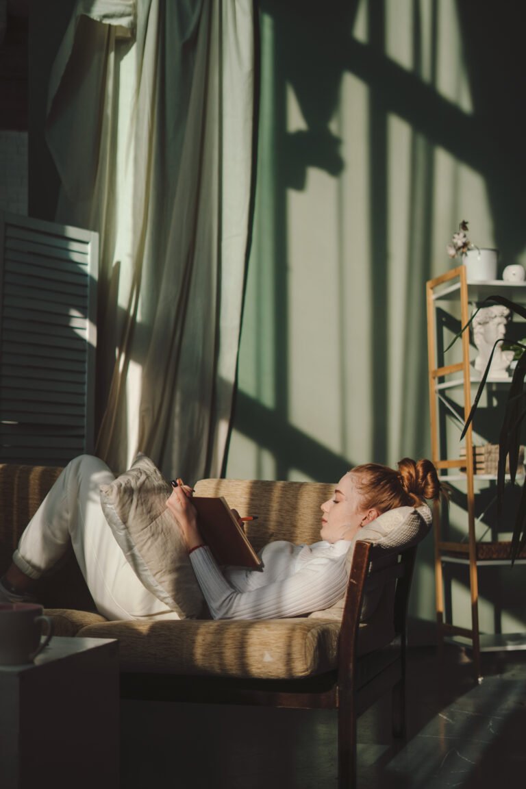 Creative writing woman making notes in diary while giving herself a facial mask lying in the morning on sofa. Relaxed female planning, enjoying free time and a calming hobby