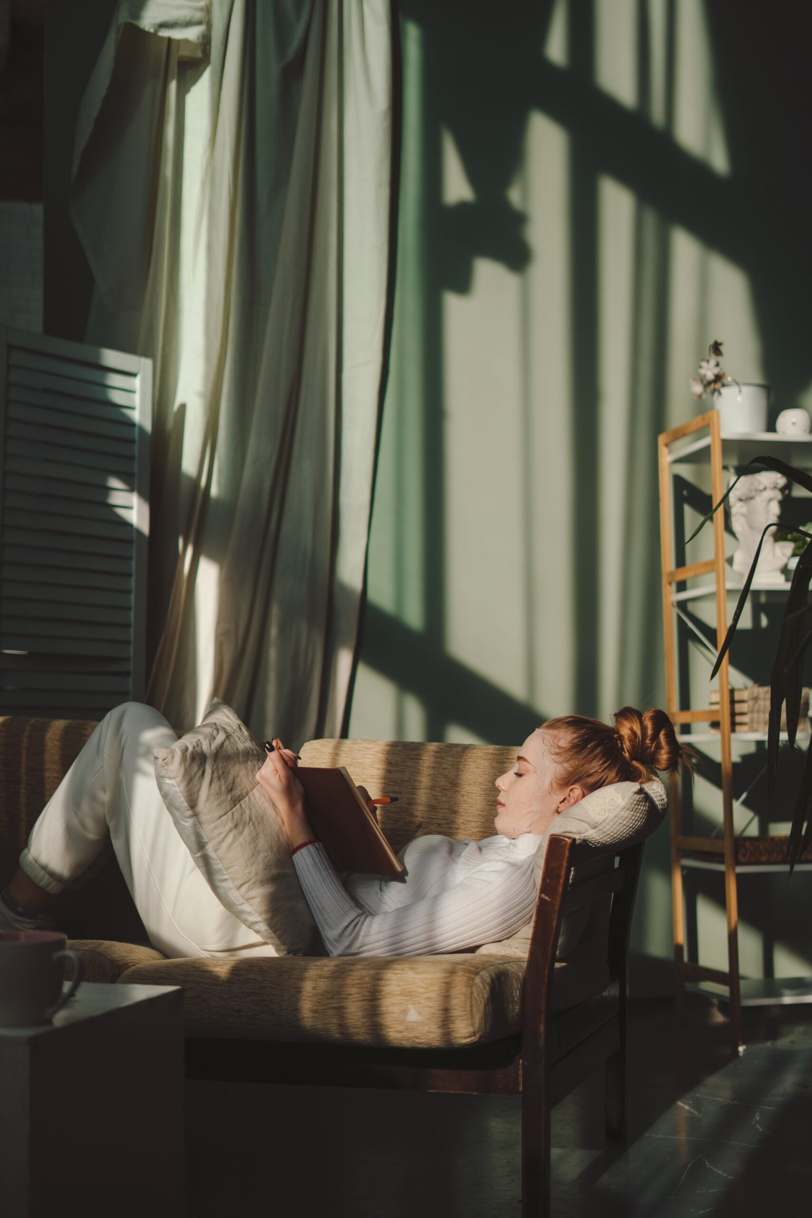 Creative writing woman making notes in diary while giving herself a facial mask lying in the morning on sofa. Relaxed female planning, enjoying free time and a calming hobby