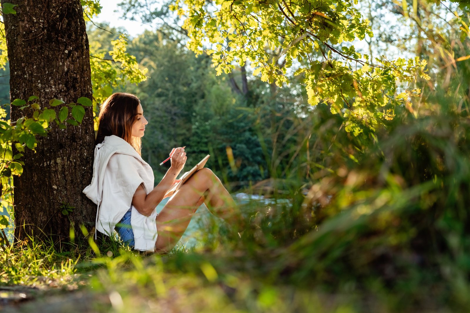 Young attractive girl writing and drawing in notebook sitting under the tree on nature in sunlight. Closeness to nature inspires creativity. Urban escapism.