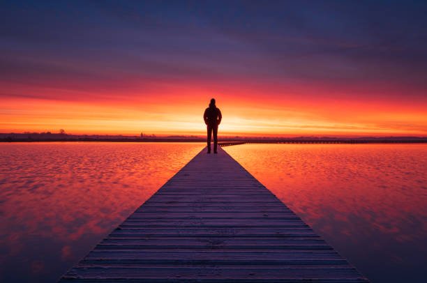 A man enjoying a colourful dawn on a small boardwalk over a lake.