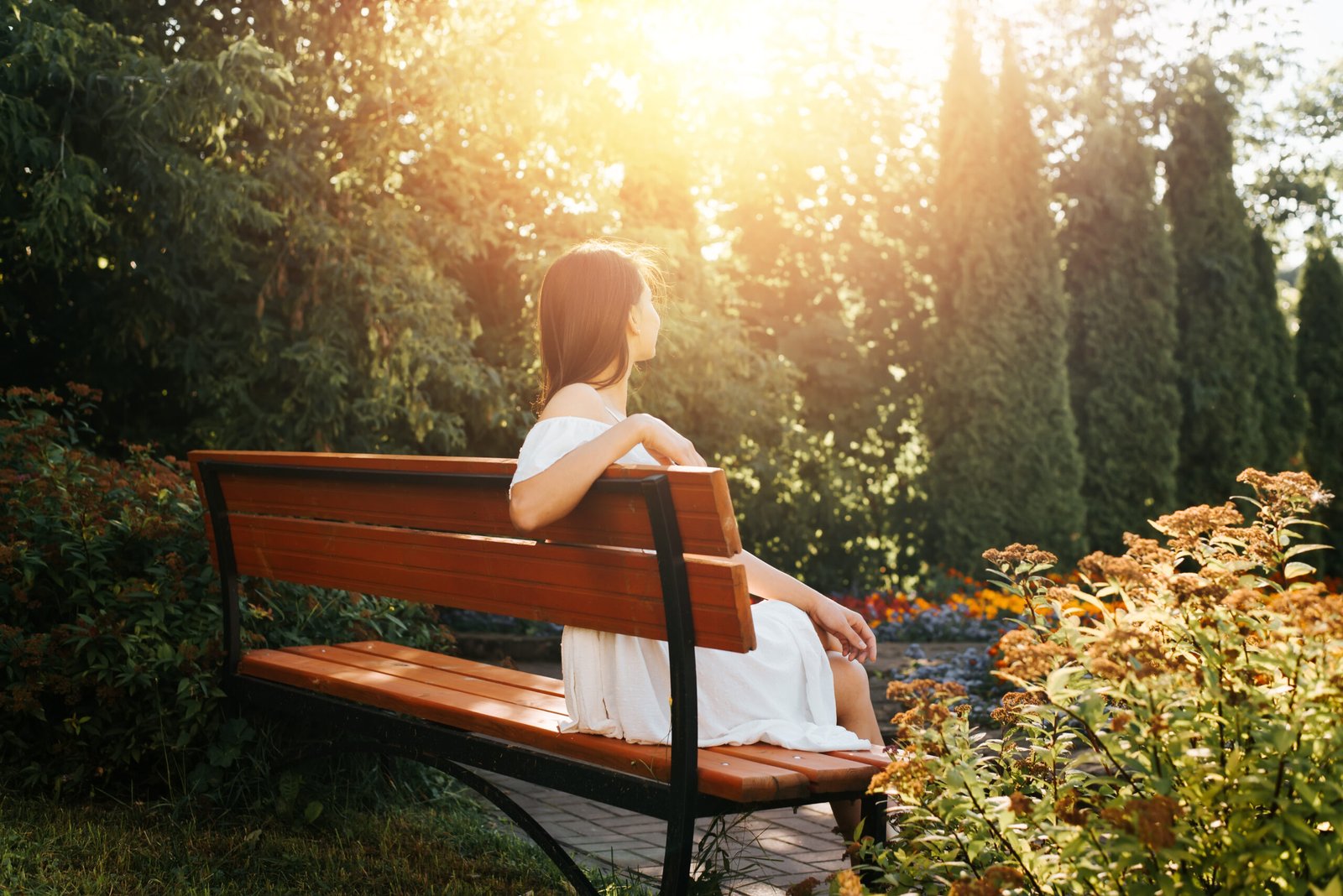 Young brunette woman in white summer dress resting, relaxing in flower garden outdoors. Back view of an elegant lady sitting on bench and enjoying sunset