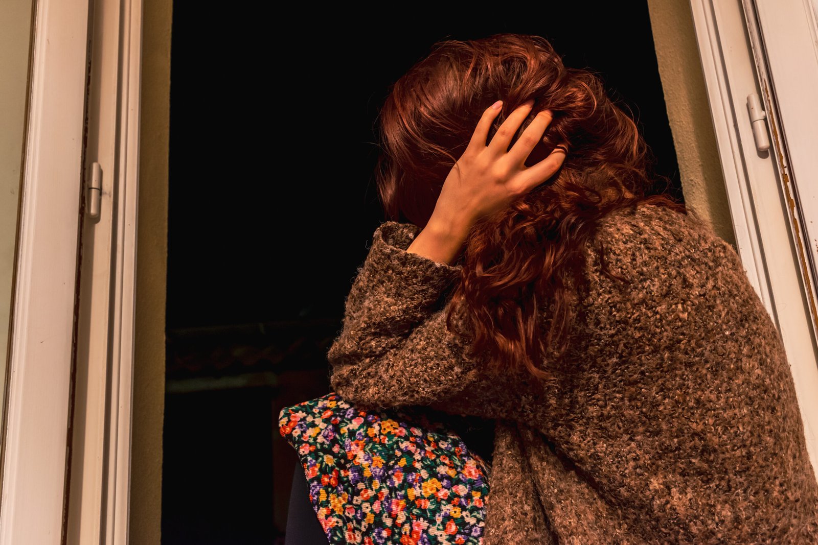 A close-up shot of an unrecognizable young redhead Caucasian woman sitting on a windowsill looking out at the street at night with her hand in the messy hair