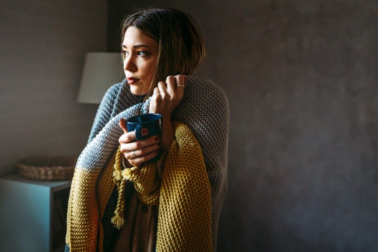 Freezing woman covered with a cozy blanket. Female wrapped in warm blanket, drinking hot drink.