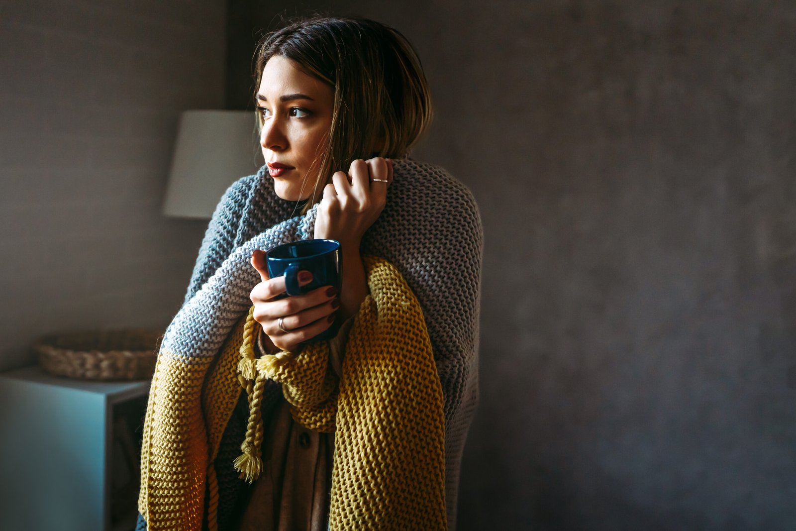 Freezing woman covered with a cozy blanket. Female wrapped in warm blanket, drinking hot drink.