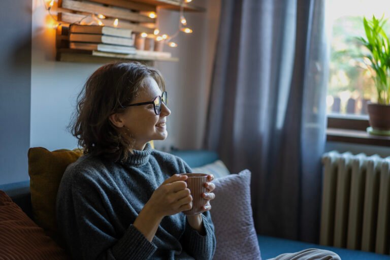 Cheerful curly young woman in warm sweater glasses enjoying a mug of tea while sitting on the sofa at home