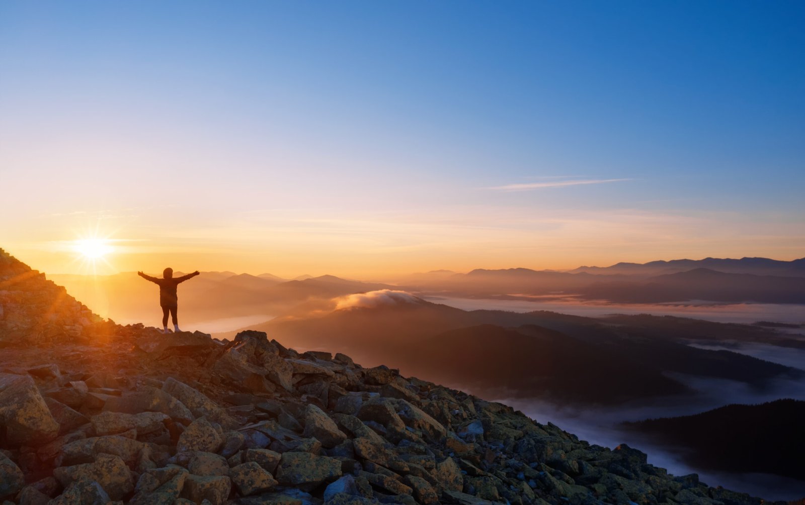 Girl on a mountain top with outstretched arms. Dramatic landscape of a lone hiker looking at mountains in fog at sunrise.