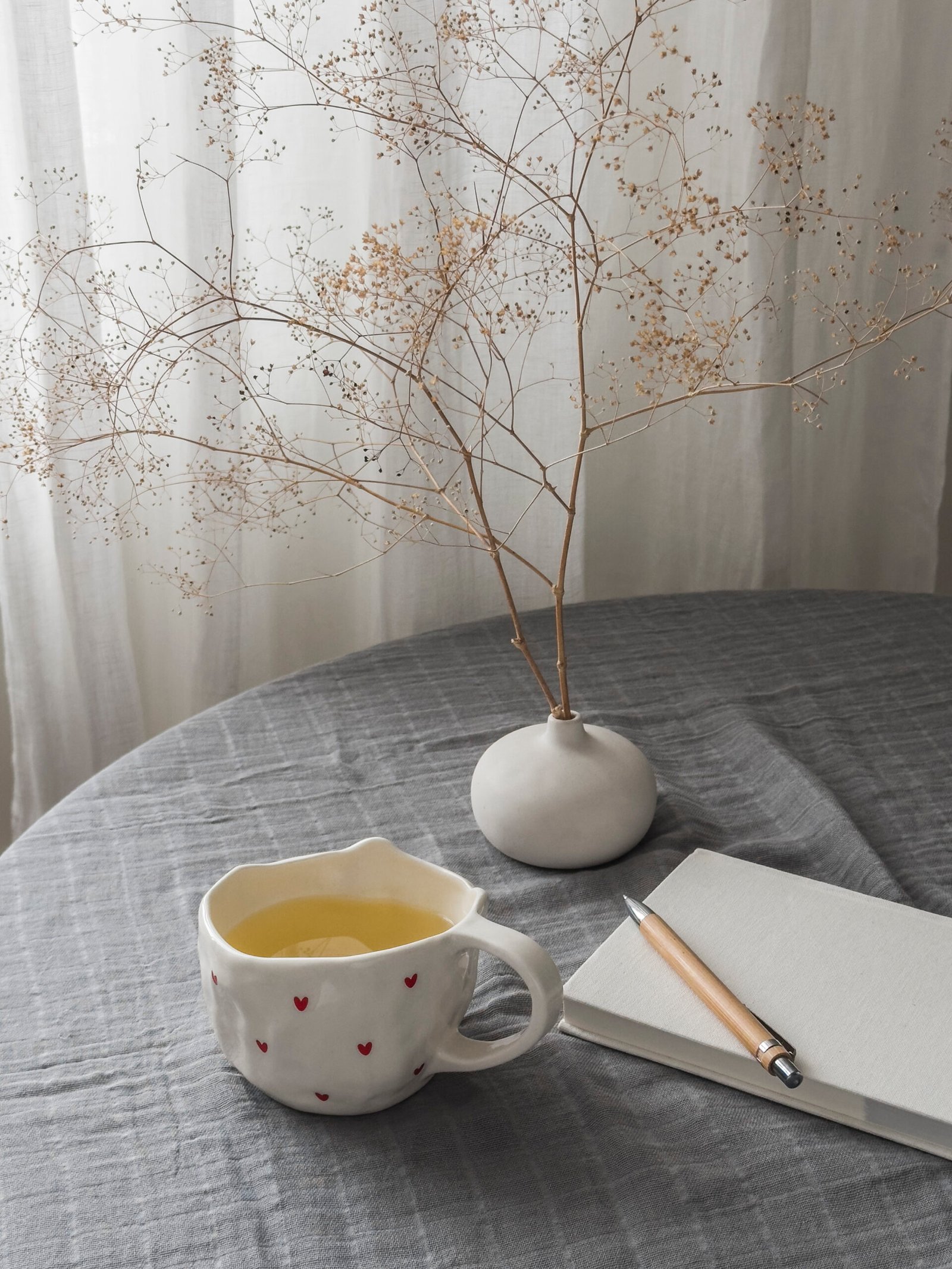 A cozy house - a cup of green tea, a notebook, a dried flower in a ceramic vase on the table