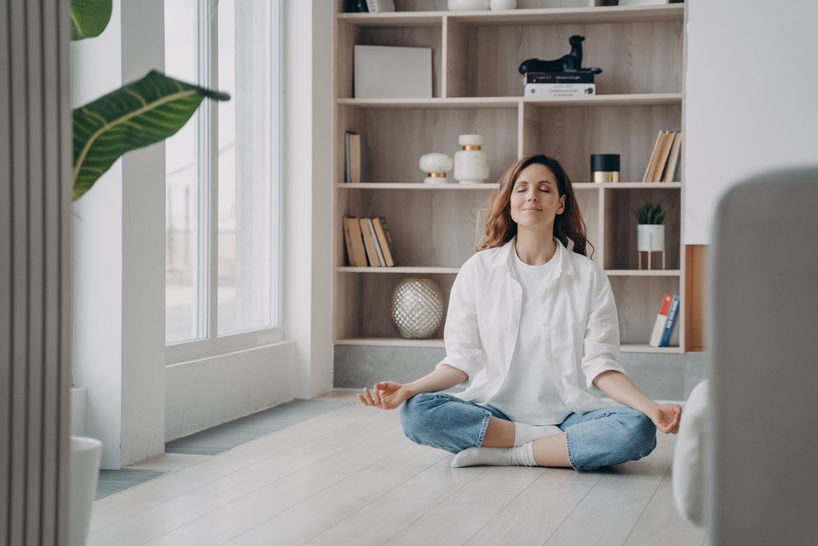 Calm happy latin woman is practicing yoga. Girl is sitting near window in morning in lotus asana.