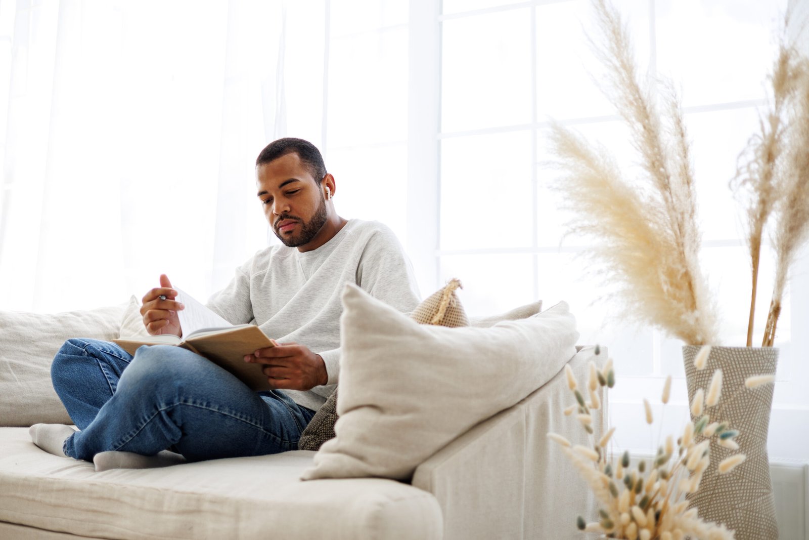 Young african american man in earphone reading book on couch at home