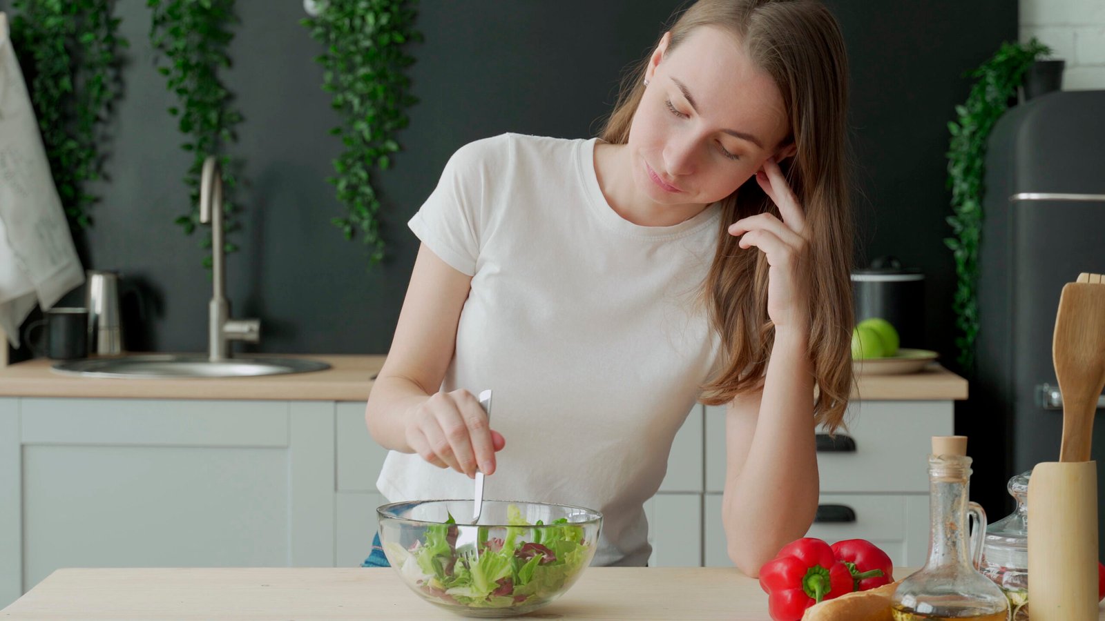 Unhappy woman eating vegetable salad at table in kitchen. Displeased young woman eating green leaf lettuce