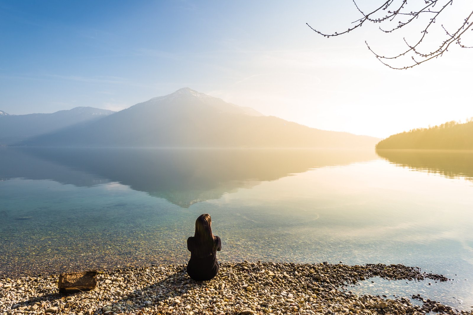 A small figure of a woman sits on the shore of a lake in the background of an epic landscape.
