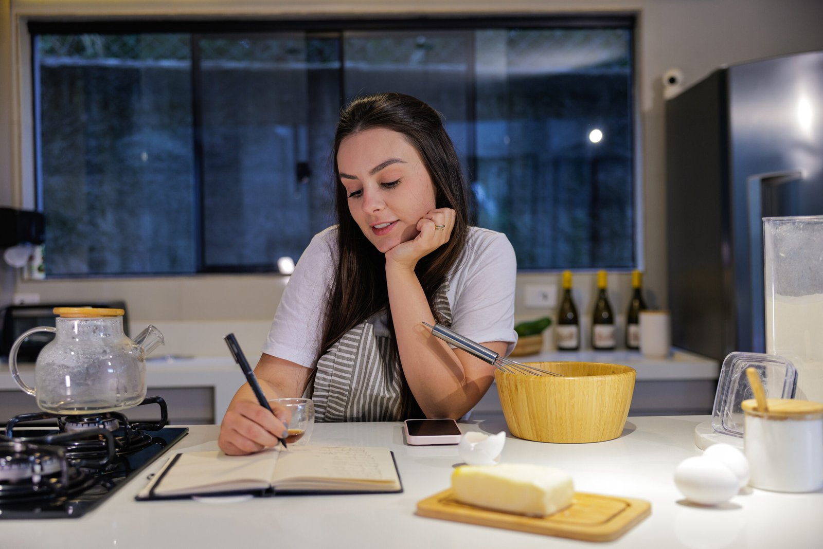 Young woman writing down recipe in notebook and drinking coffee in kitchen