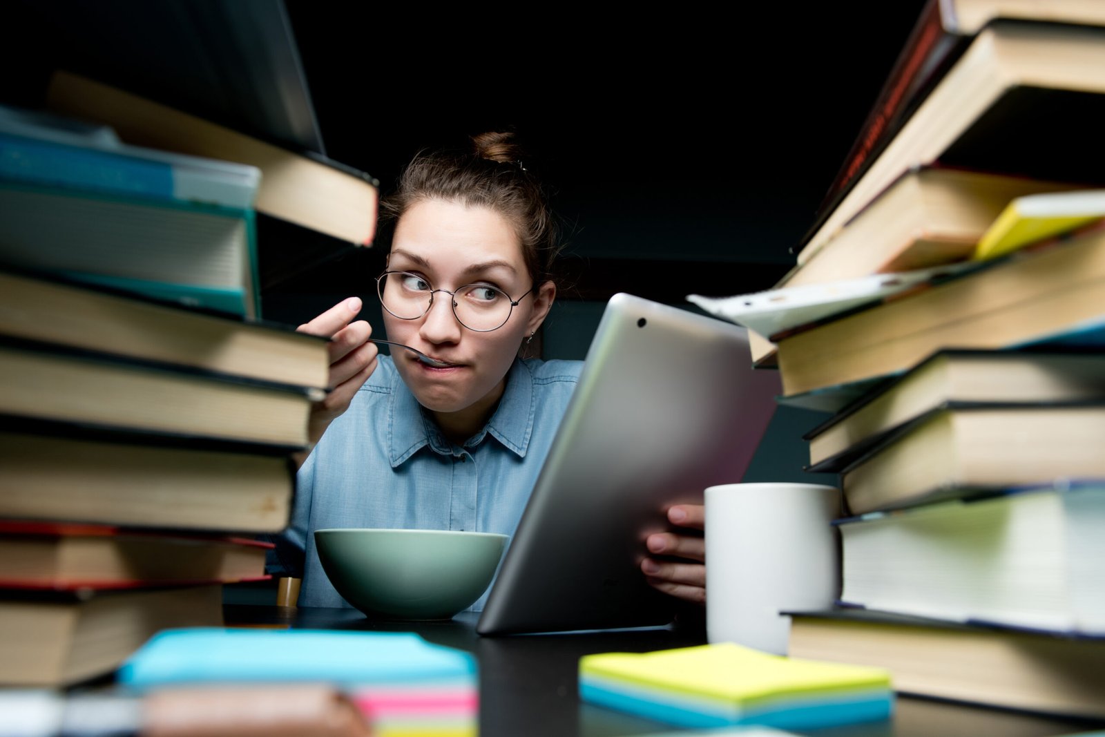 A young girl is eating and looking at a modified tablet. a lot of books on the table