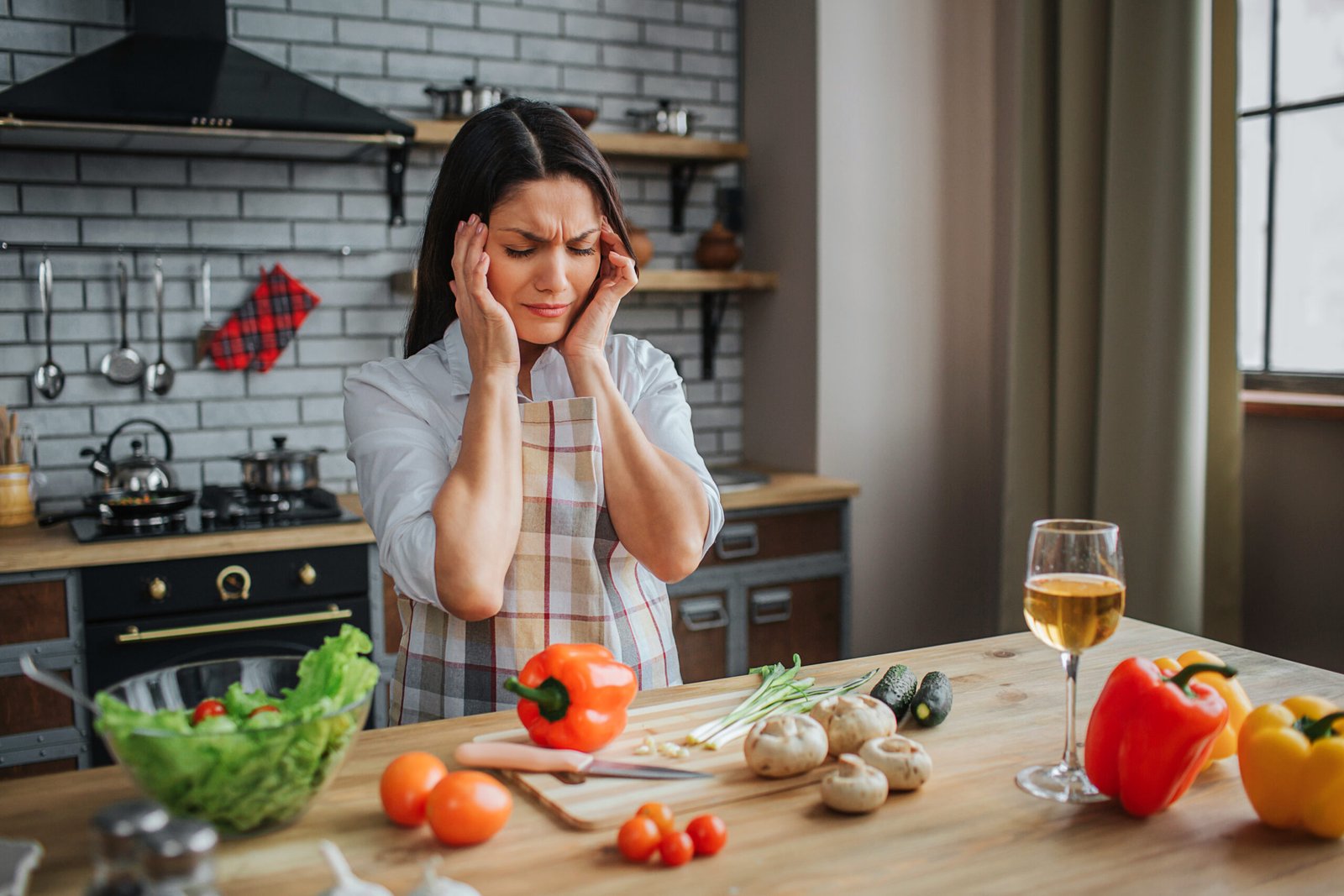 Young sick woman has headache. She hold hands on head. She sit in kitchen at table. Woman feel bad. Vegetables lying on table.