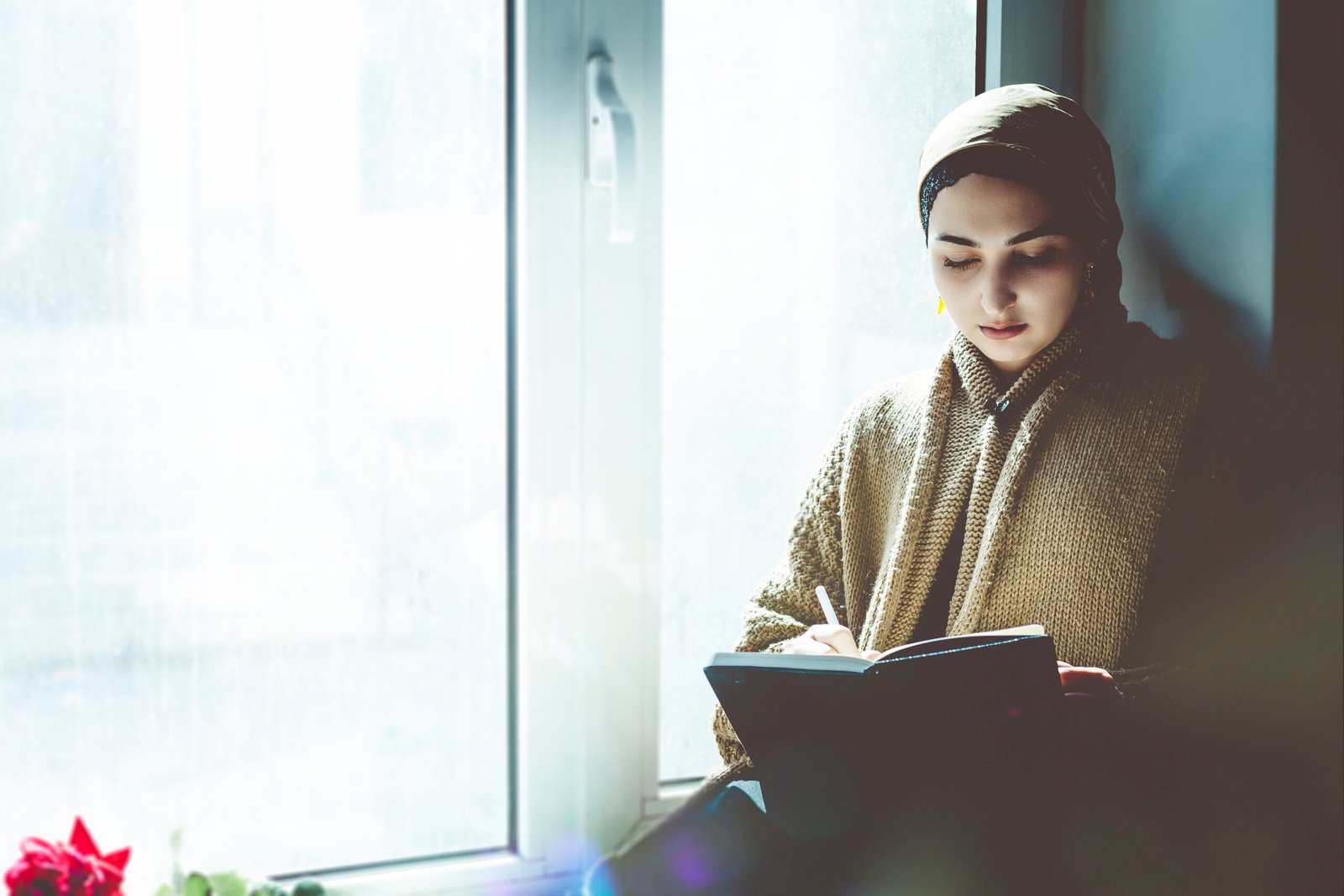 Young Muslim woman sits on the window sill. Middle-eastern writer woman sitting thoughtfully near the window and writing her story on the notepad. Iranian female making notes on her diary