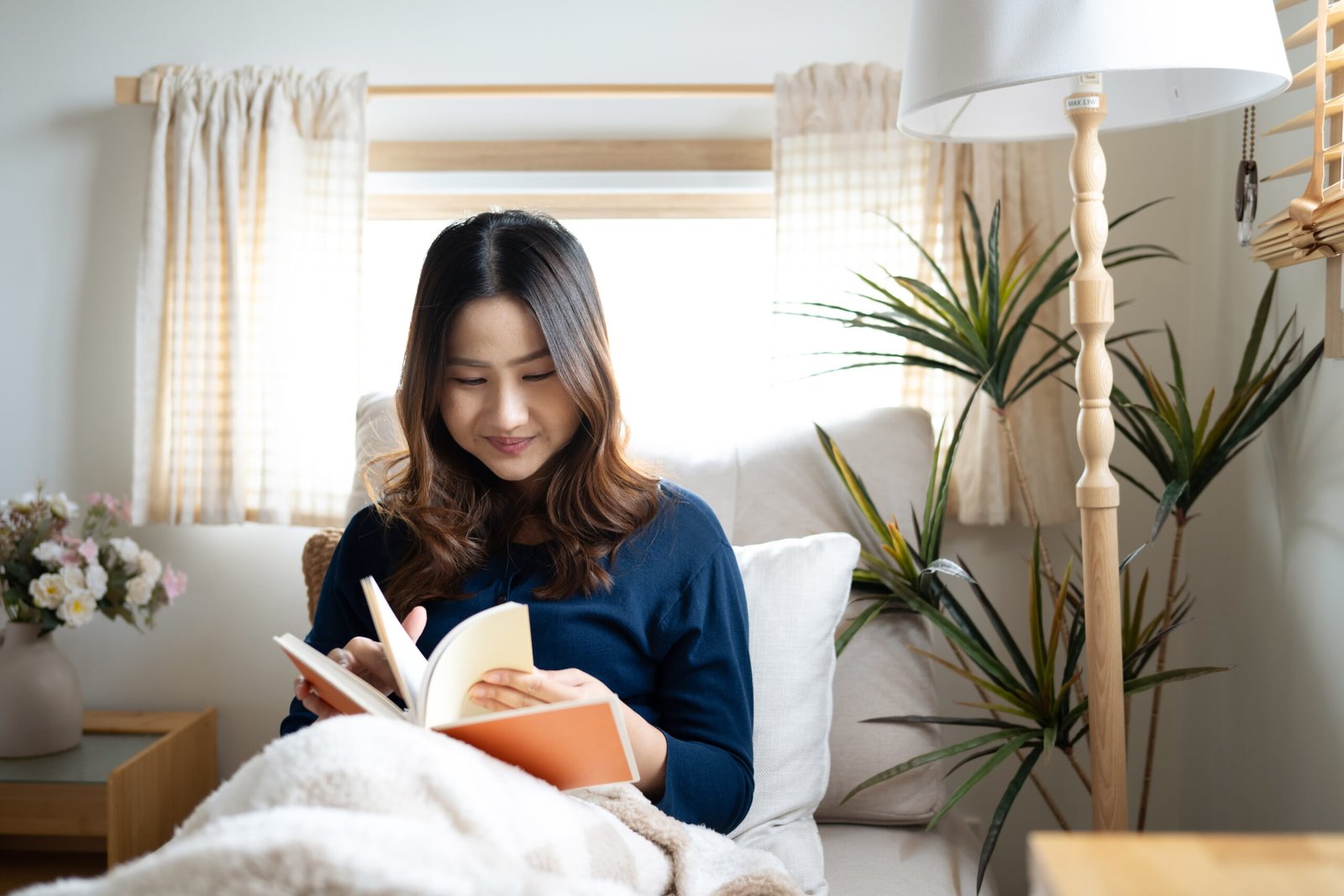 Calm young woman sitting under soft blanket reading a book by the window at home
