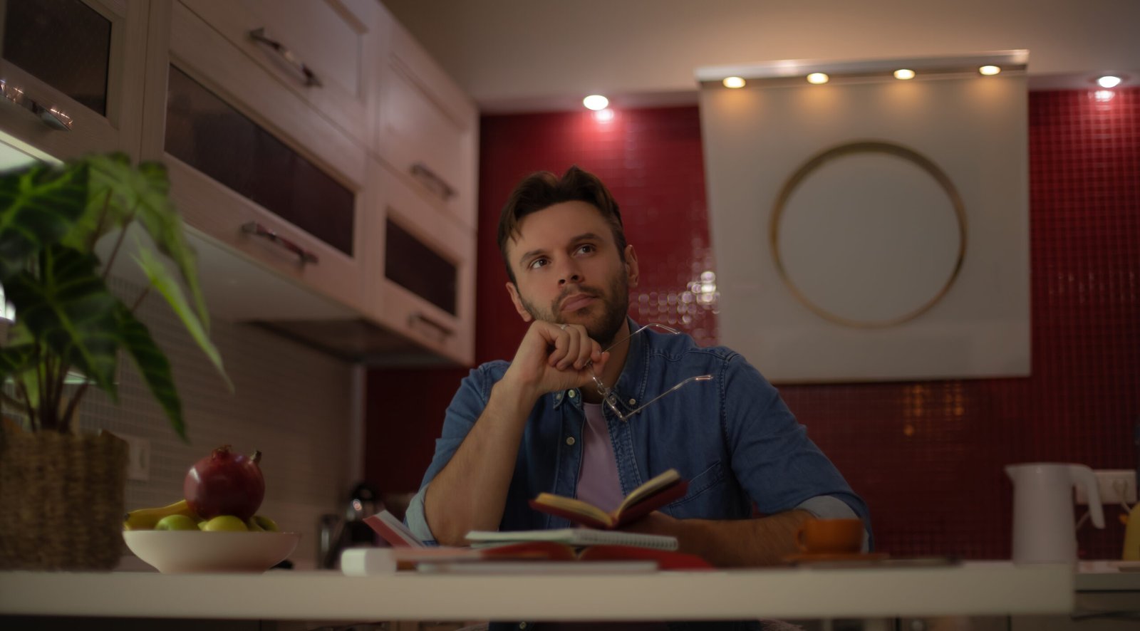 Thoughtful young man sitting at a kitchen table with books, notebook, and coffee, holding his glasses and looking away, appearing deep in thought or brainstorming ideas.