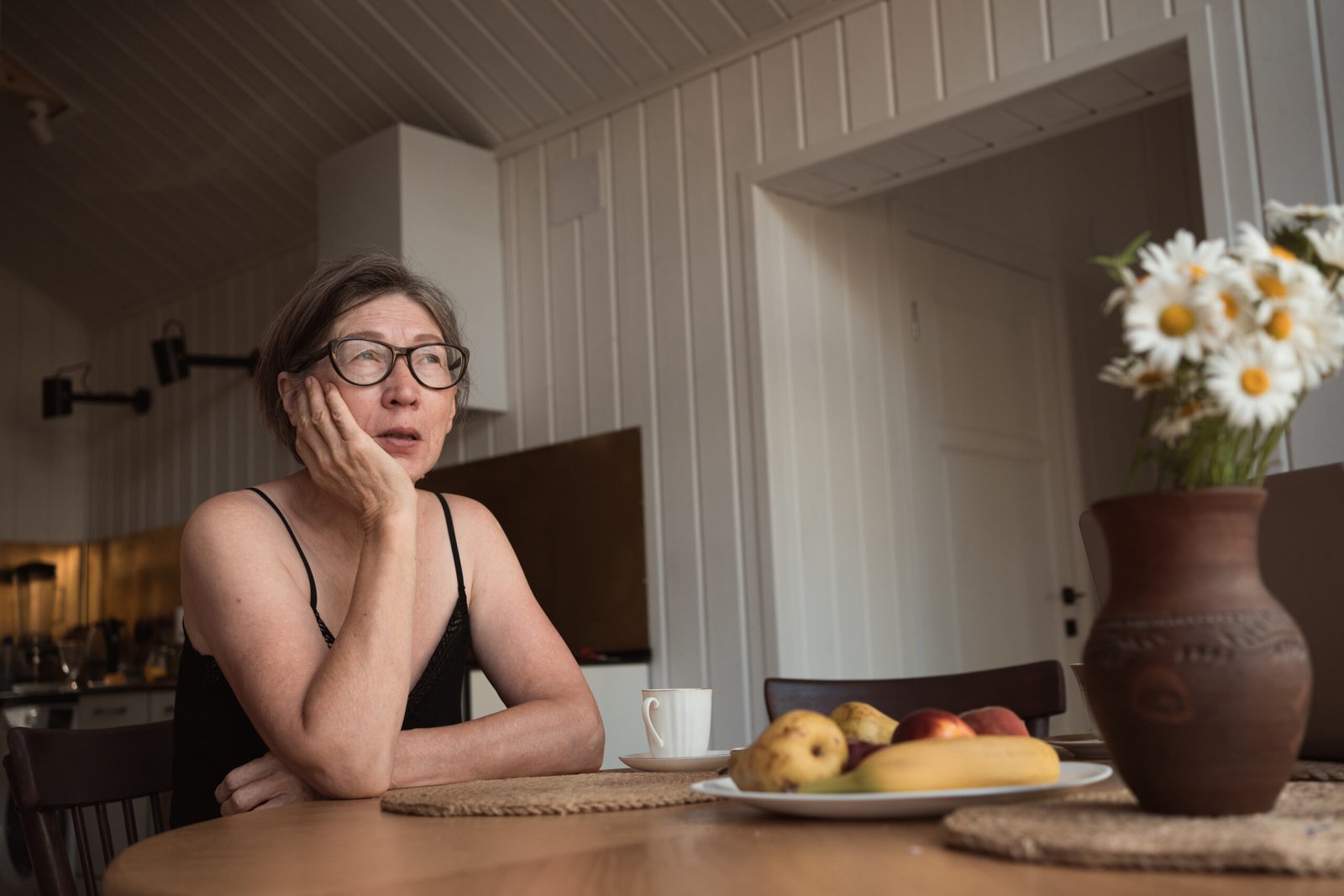 Worried senior 60s woman wearing glasses sitting at the kitchen table