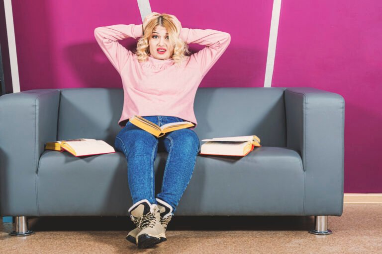 Girl blonde sits on sofa. There are open books around her. She is panicking and holding head with hands.