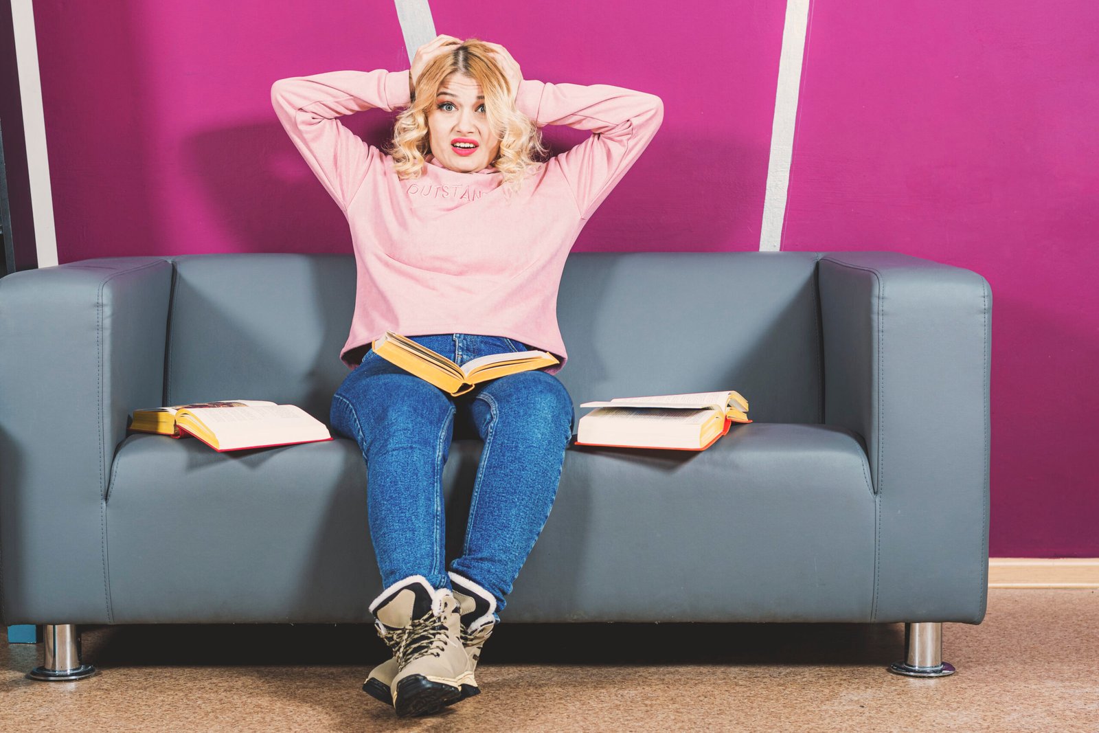 Girl blonde sits on sofa. There are open books around her. She is panicking and holding head with hands.