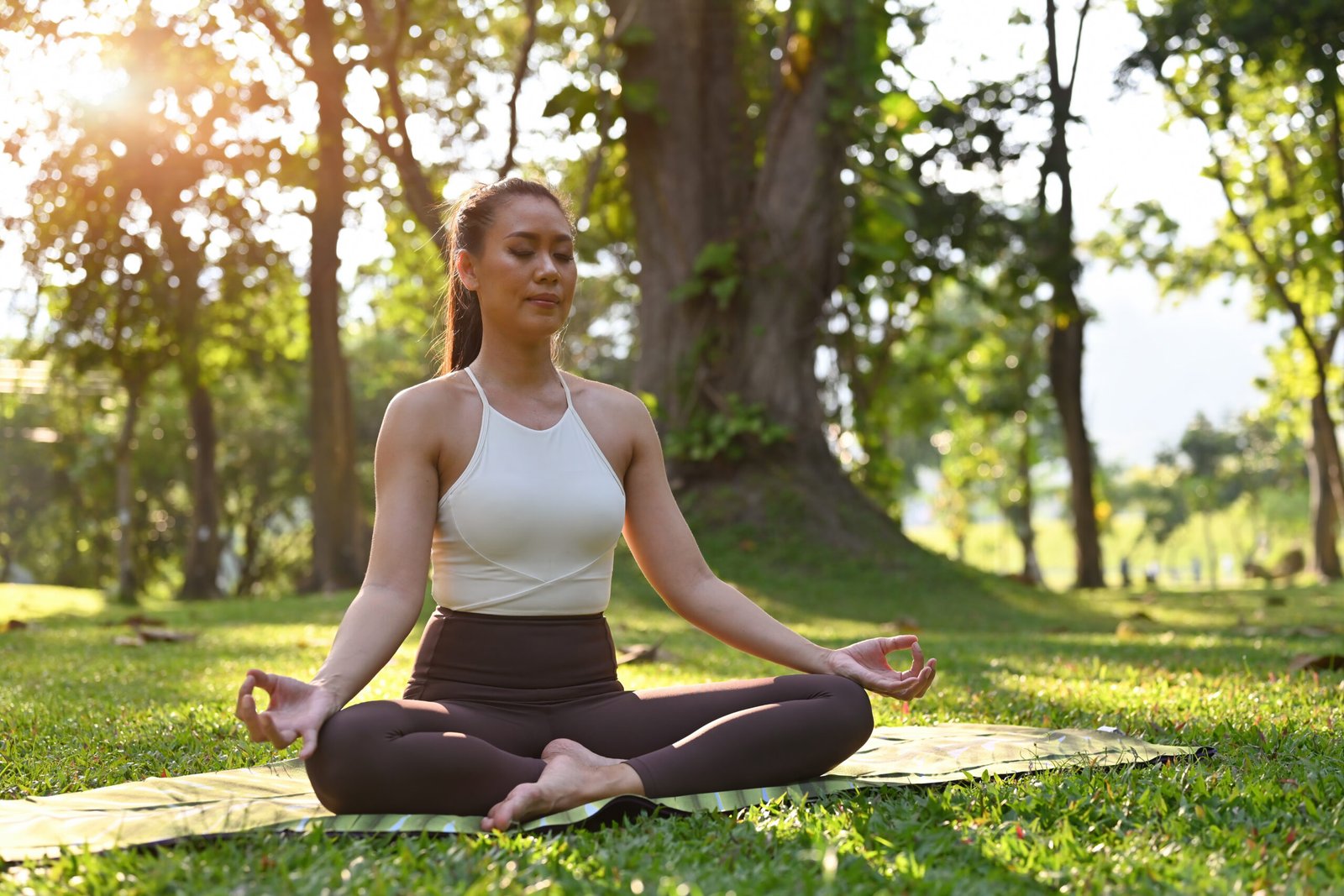 A woman practices meditation in the lotus position, her hands in a relaxed mudra, surrounded by vibrant greenery and bathed in warm sunlight