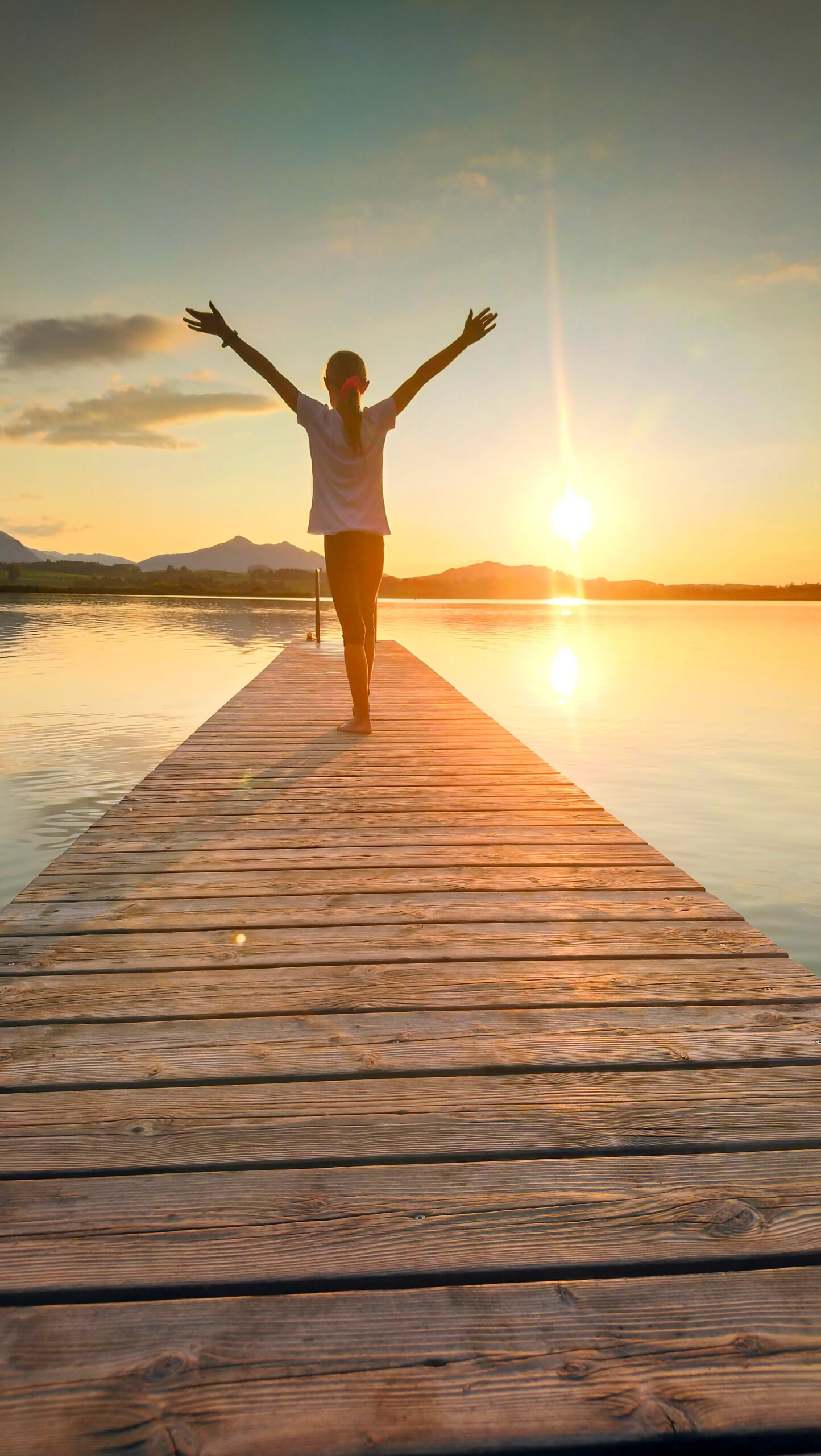 Rear View Of Woman With Arms Outstretched Standing On Pier Over Lake During Sunset
