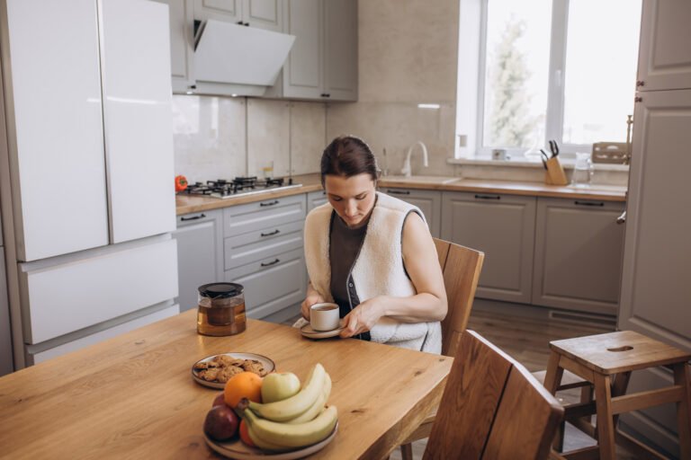 A beautiful brunette woman is having breakfast at the kitchen table with a cup of tea