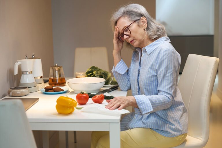 Sad gray-haired lady in casual clothes sits akitchen table