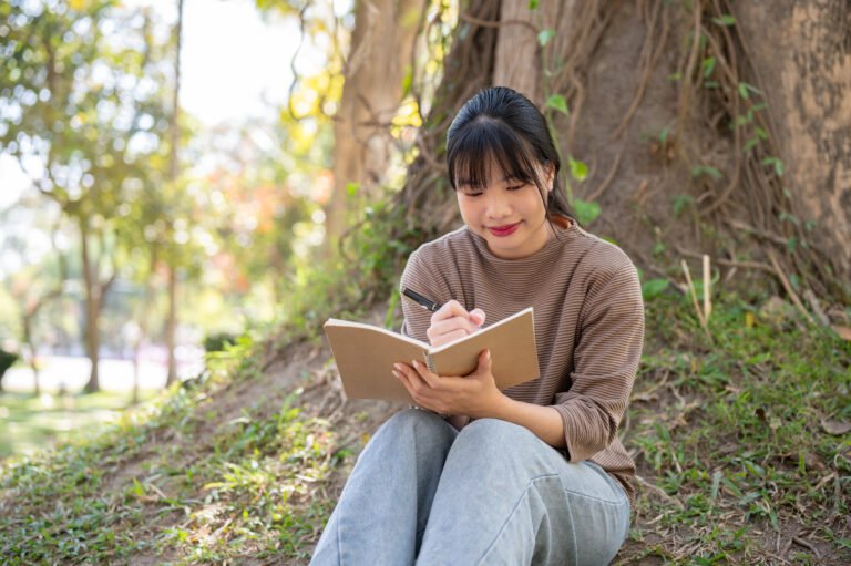 "A positive Asian woman is jotting down her ideas in a book while sitting under a tree in a park.